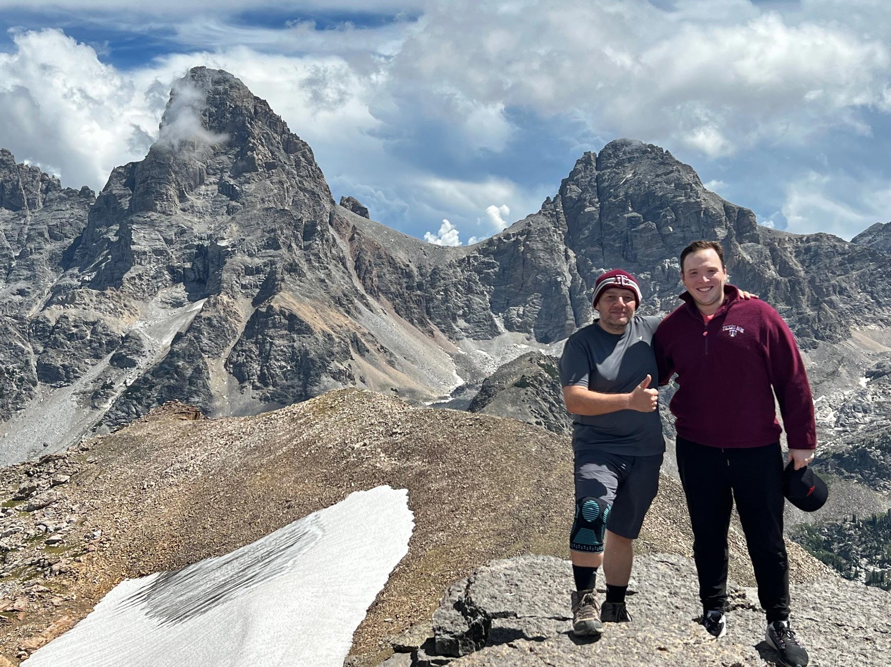 Two hikers posing in mountainous terrain with jagged peaks and clouds in the background and patches of snow on the ground. 