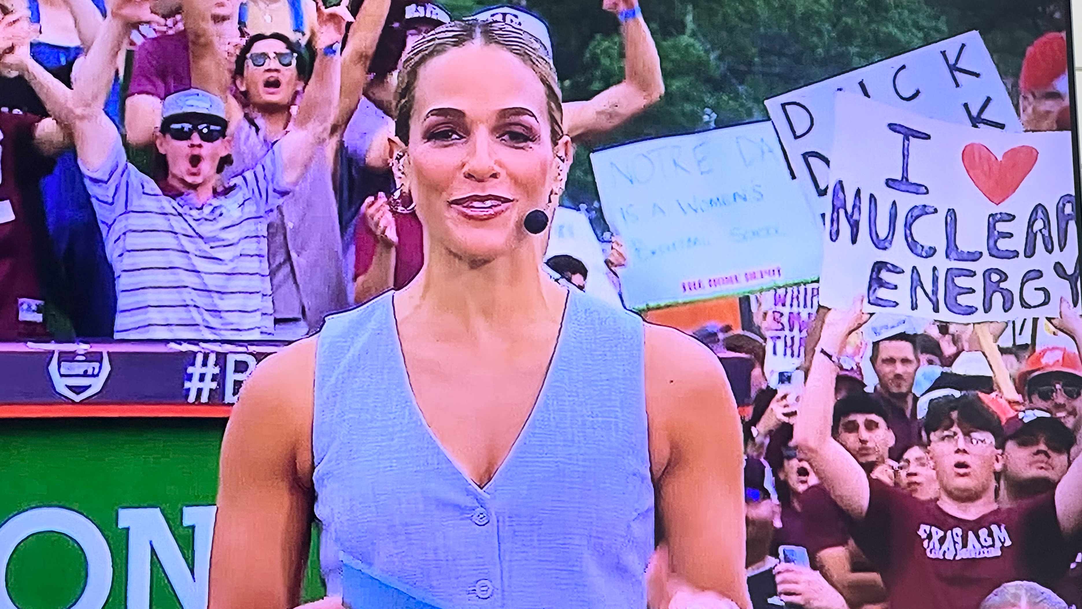  A sports reporter stands in the foreground with a microphone, smiling as she addresses the camera. In the background, a man holds up the sign I heart Nuclear Energy.