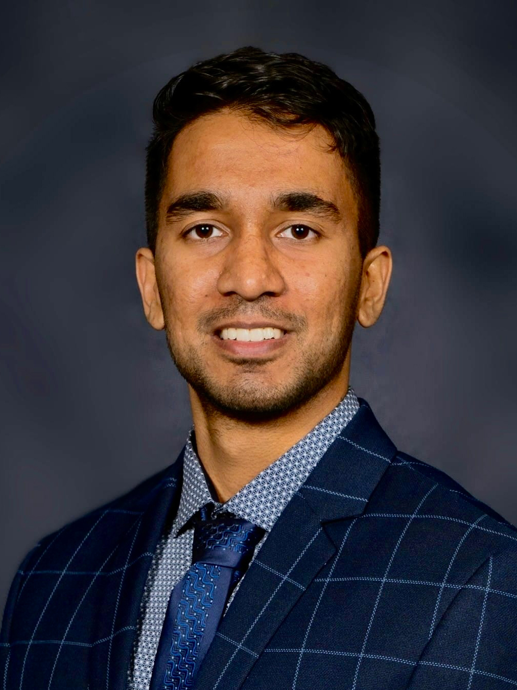 A man’s headshot wearing a dark checkered suit jacket over a patterned shirt and a tie.