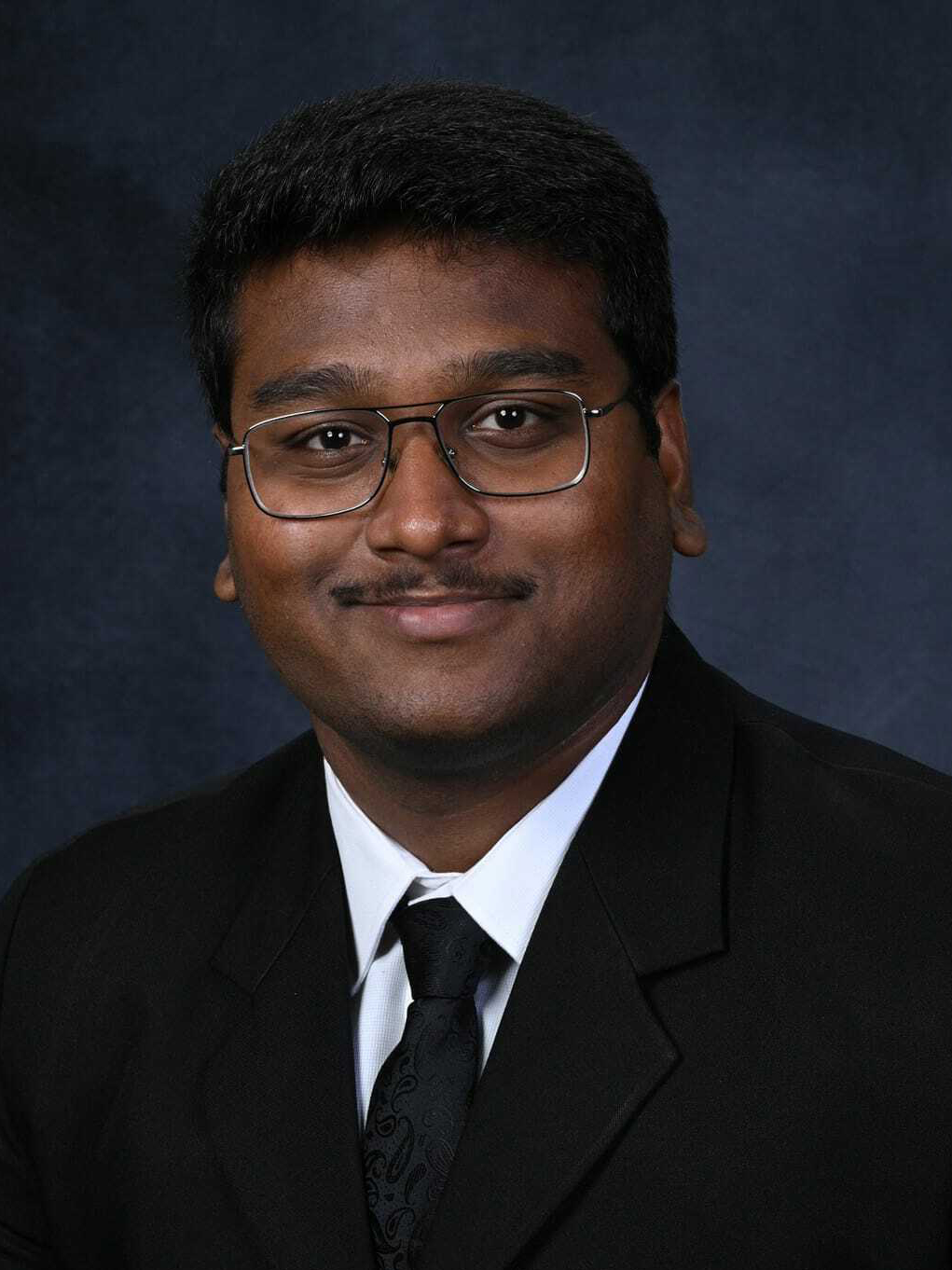 Headshot of a young man wearing a black suit and glasses smiling at the camera. 