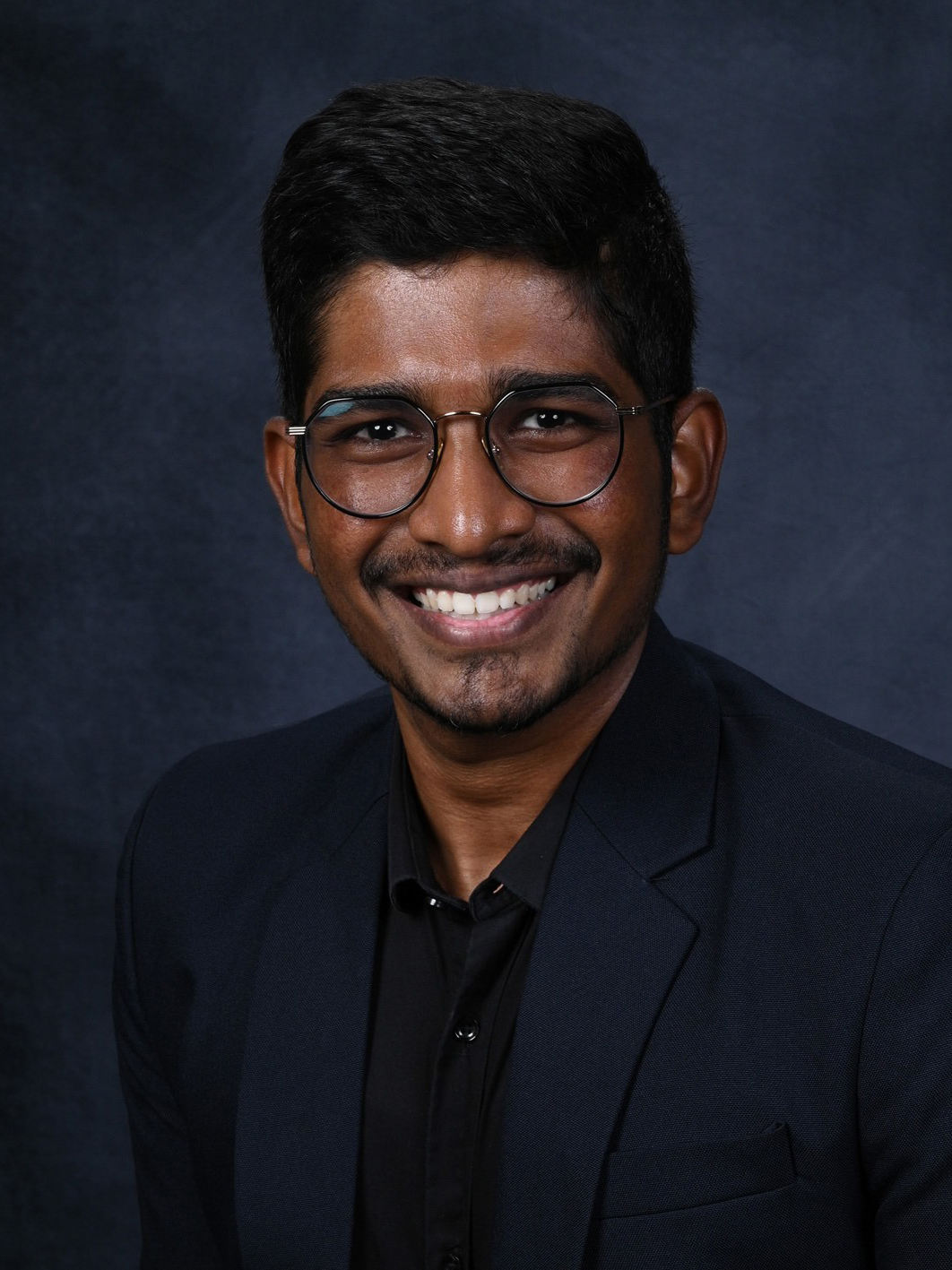 A young man smiling, wearing round glasses and a dark suit.