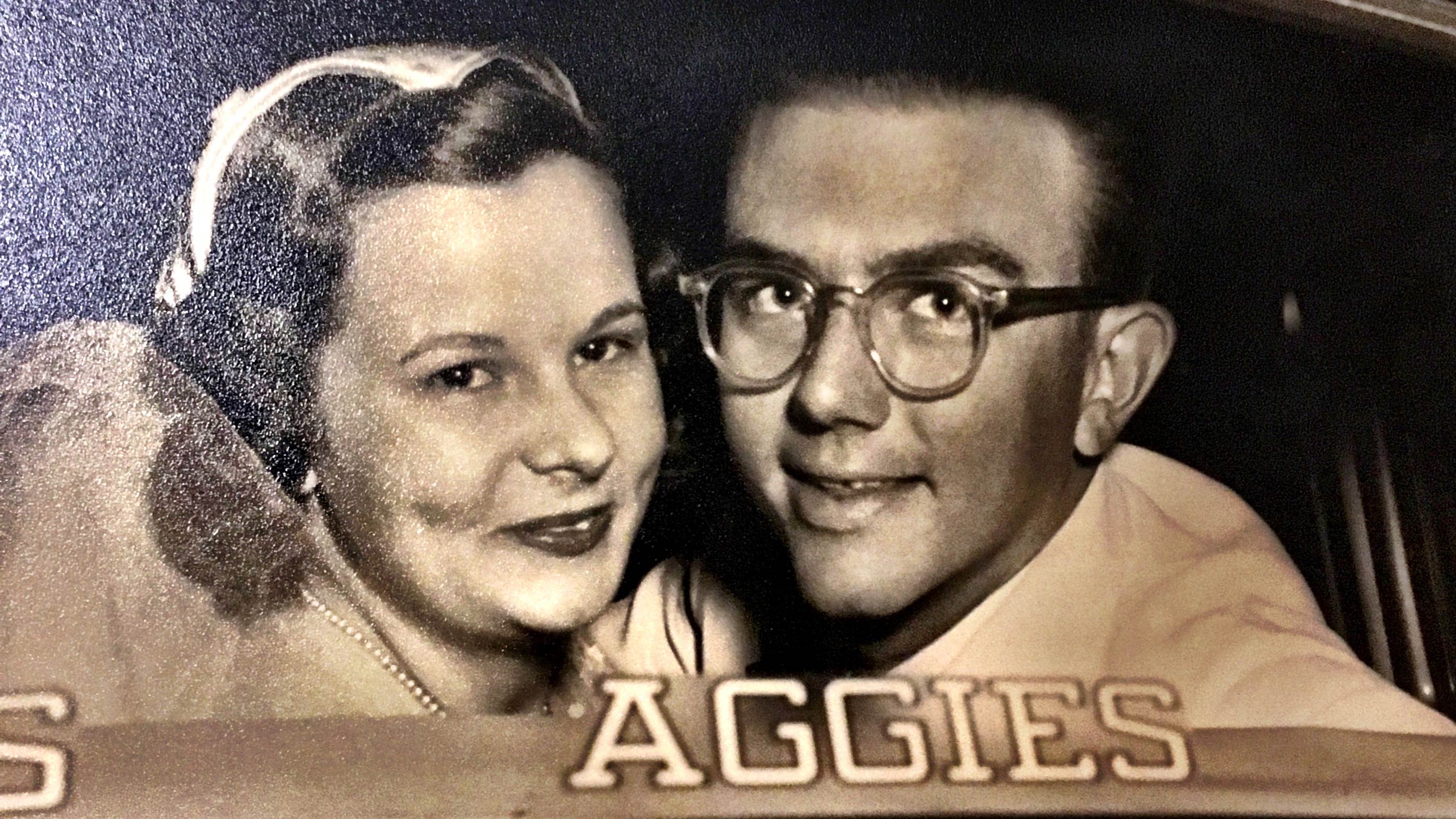 A vintage black-and-white photograph of a couple smiling at the camera from inside a vintage car. 