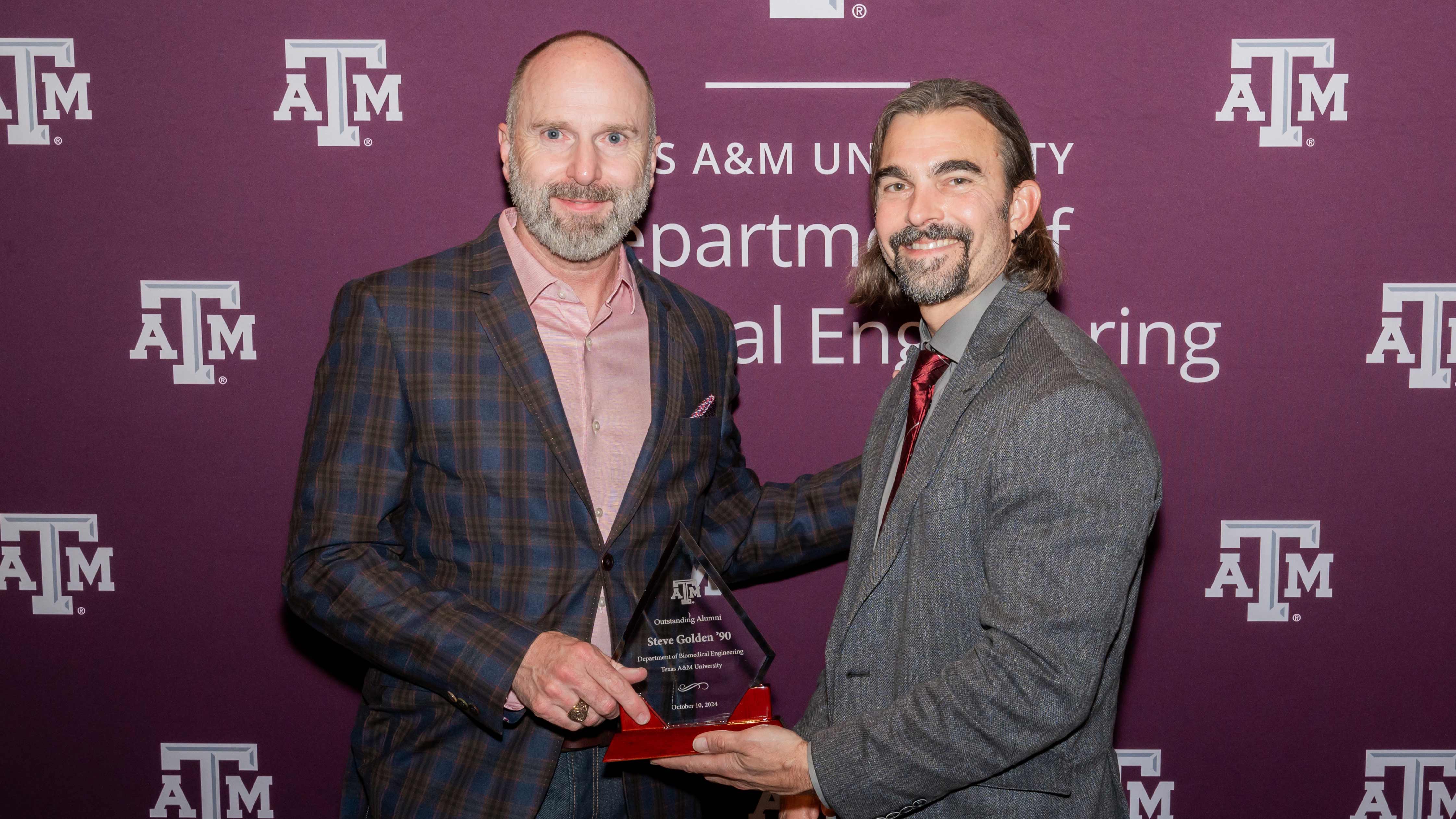 Two people in front of an A&M backdrop holding an award.