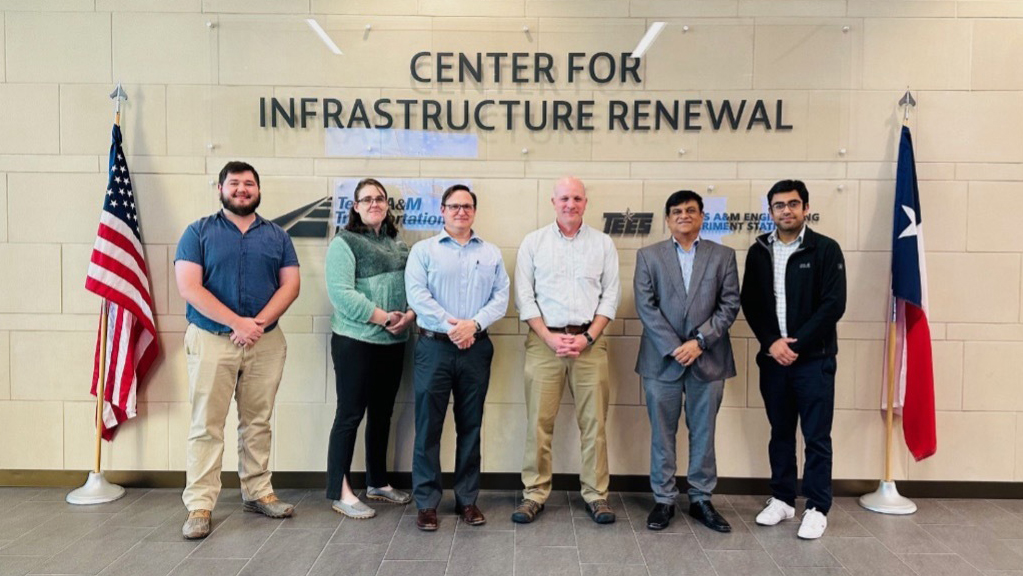 Six meeting attendees standing in front of a wall with a sign.