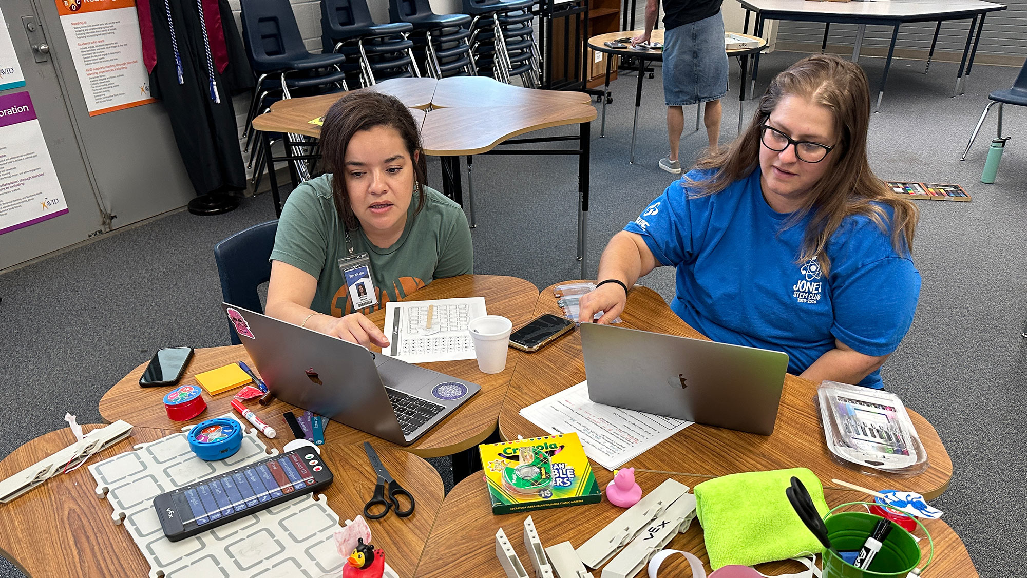 Two women sit at a student desk with laptops, art supplies and coding equipment surrounding them. 