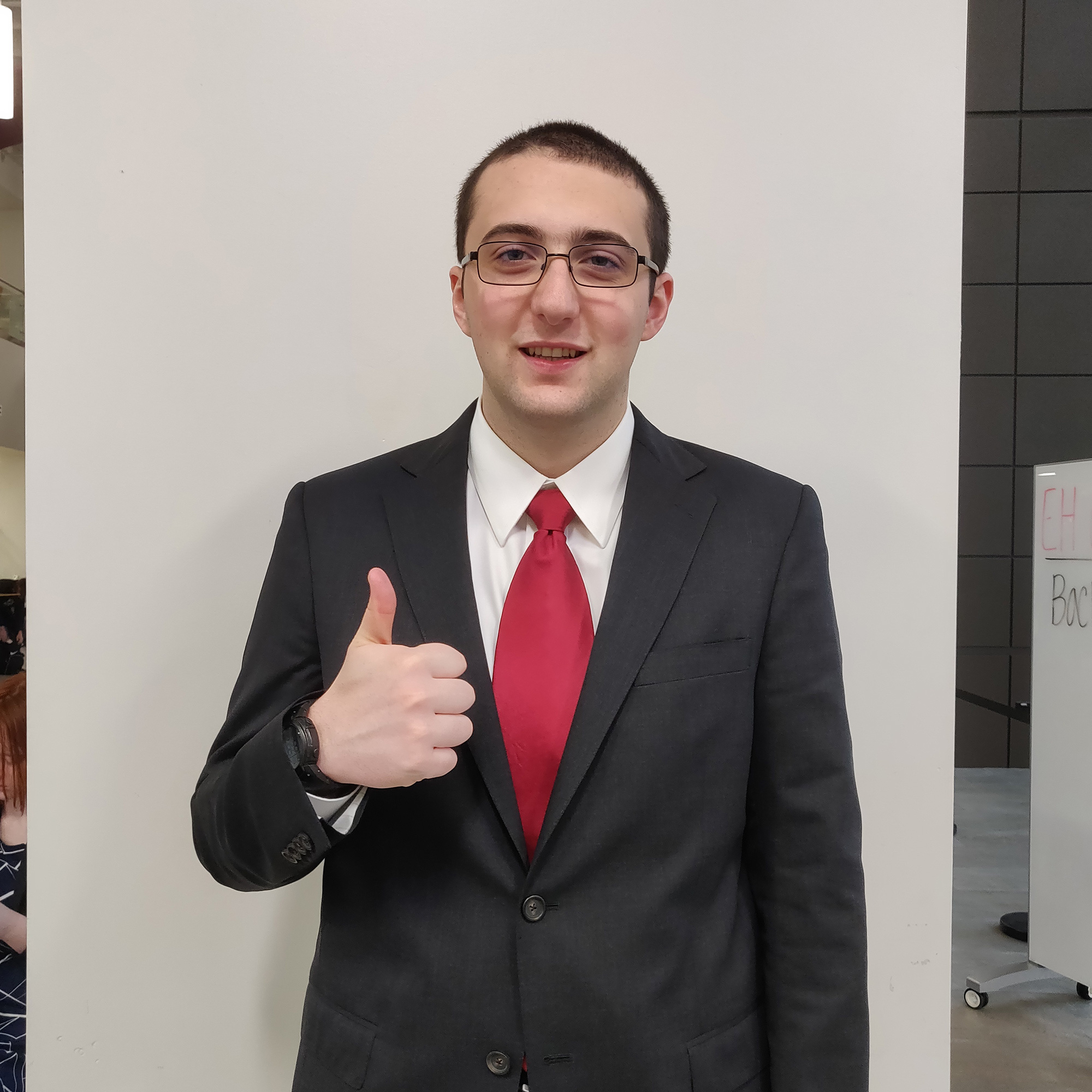 A man standing in a suit, holding up a thumb’s up.