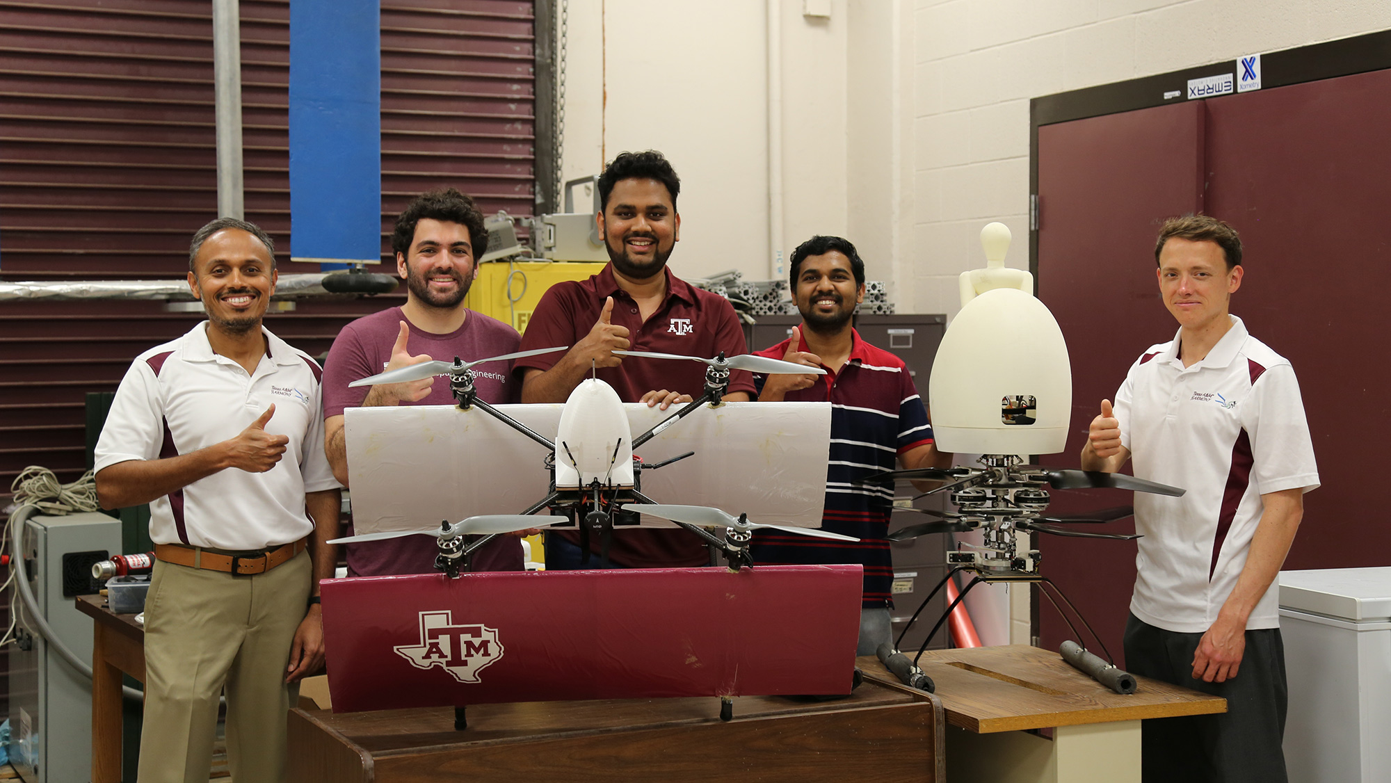 Five individuals, dressed in Texas A&amp;M shirts, stand alongside a drone in a lab setting. 