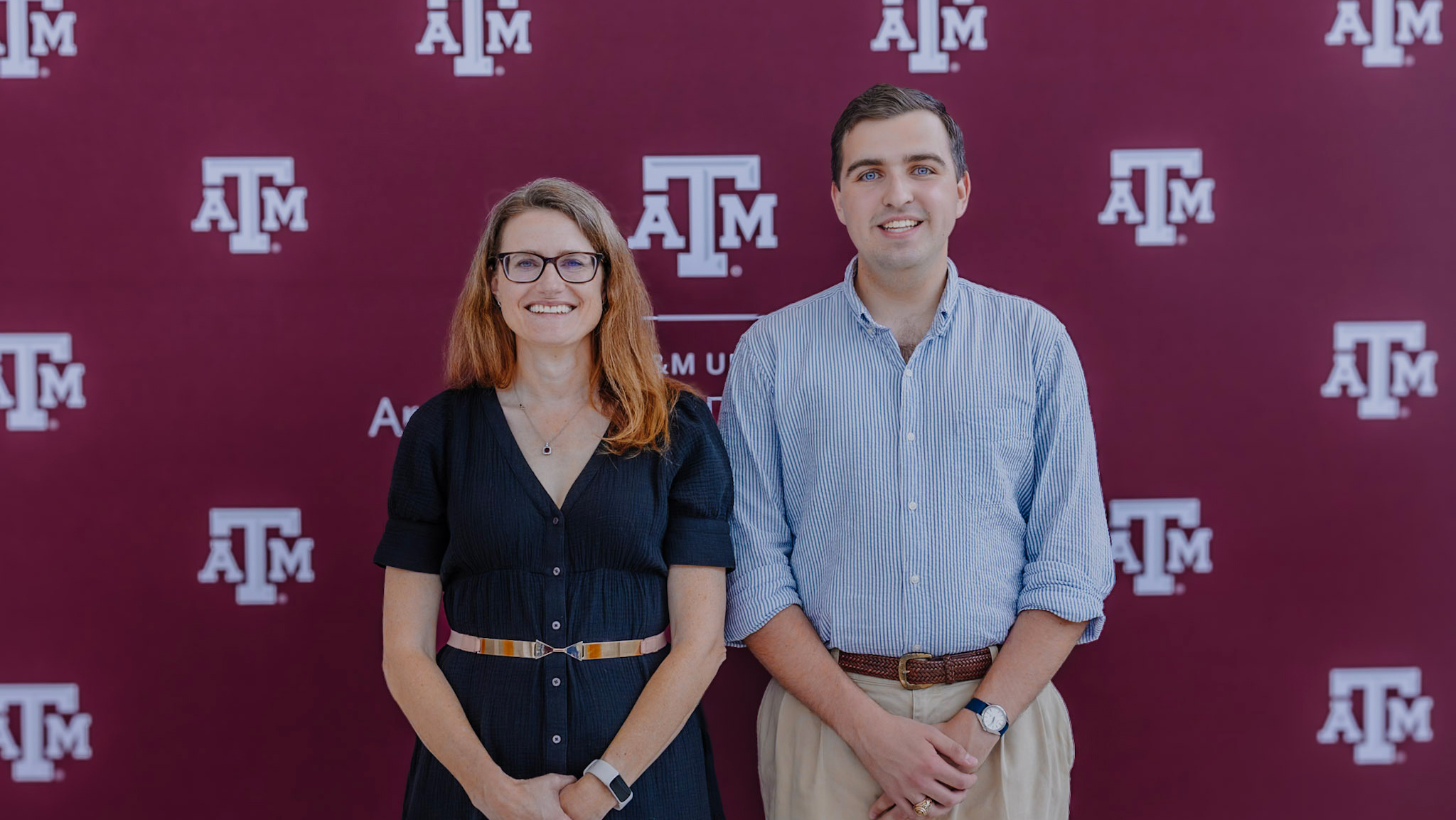 Two individuals stand in front of a maroon backdrop featuring the Texas A&M logo. 