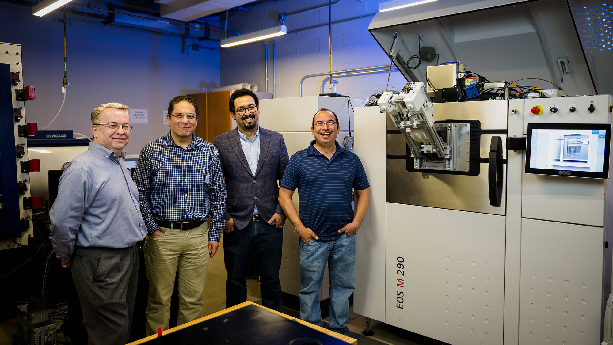 Four men pose and smile in front of a large industrial machine inside a laboratory.