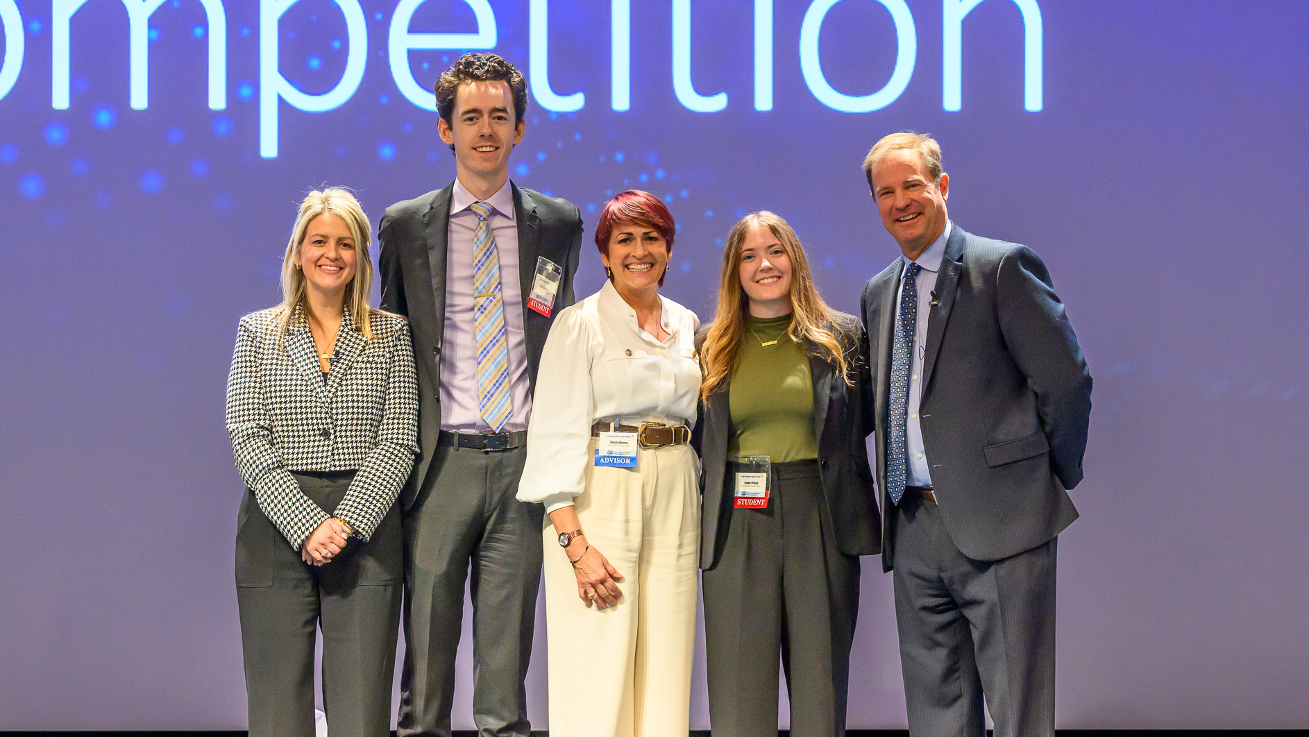  Five professionally dressed individuals stand smiling on stage in front of a large screen displaying the word Competition. 
