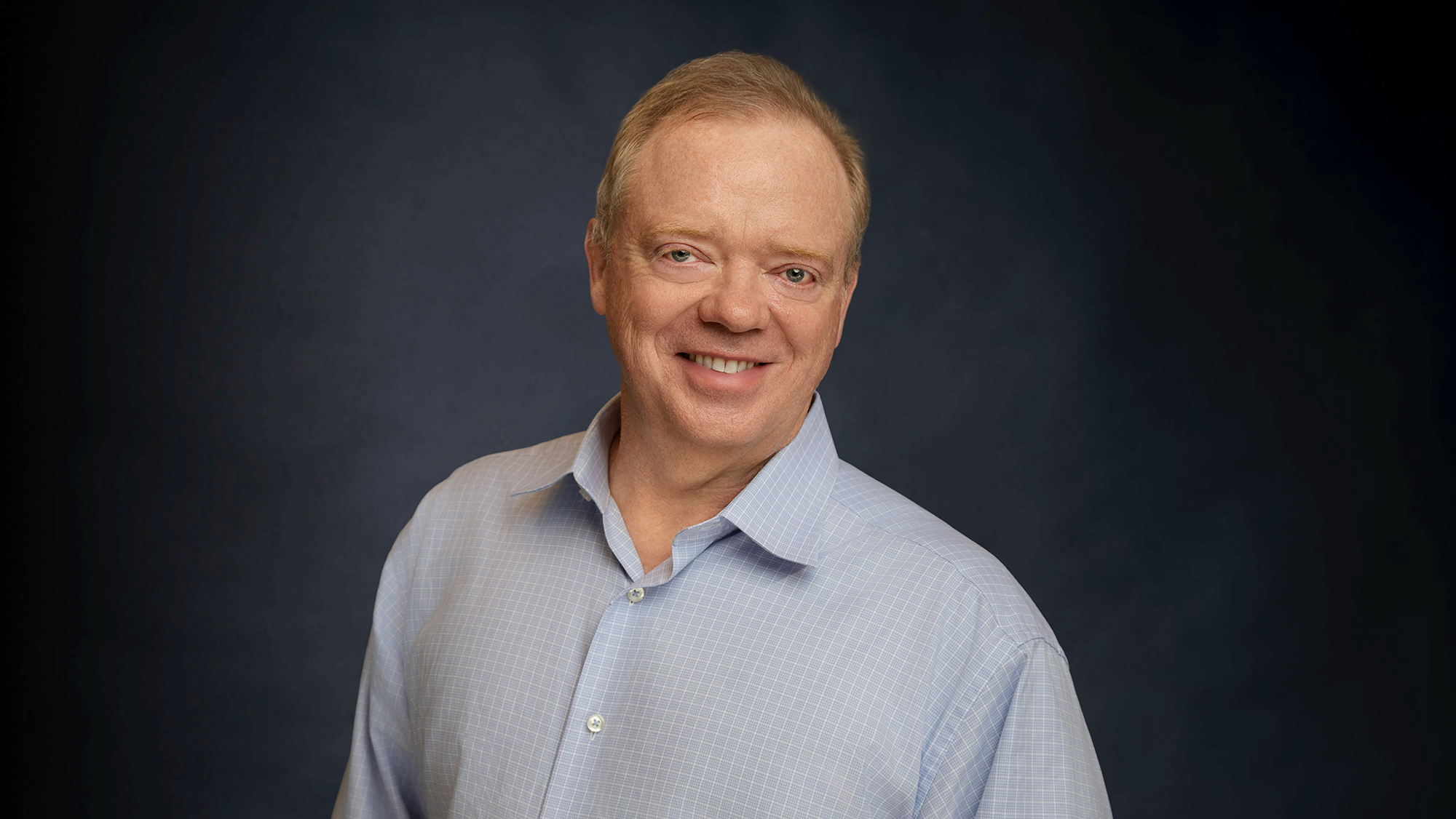 A man standing against a dark blue background, smiling.