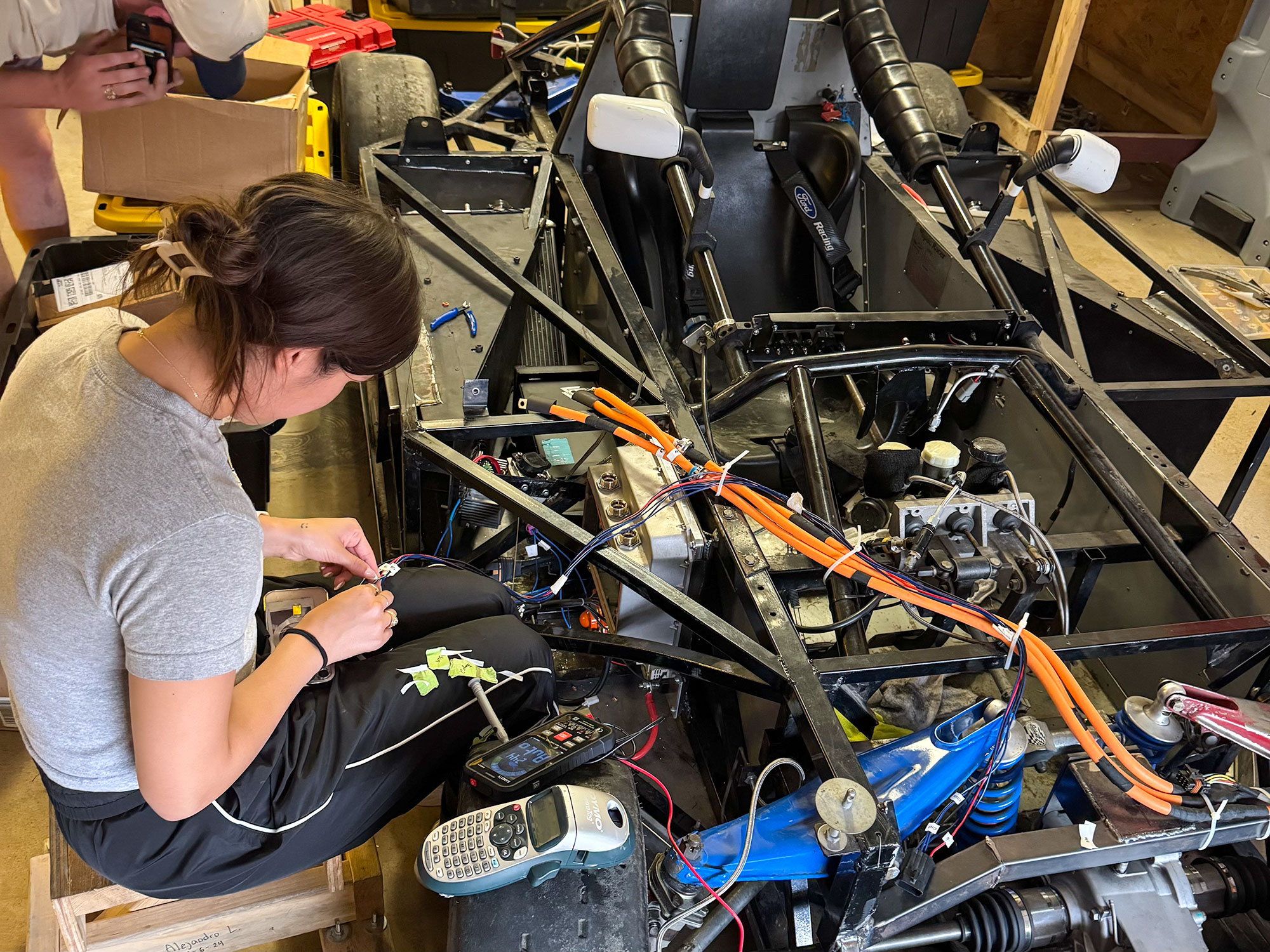 A person sits on a stool, focused on wiring inside a partially assembled race car.