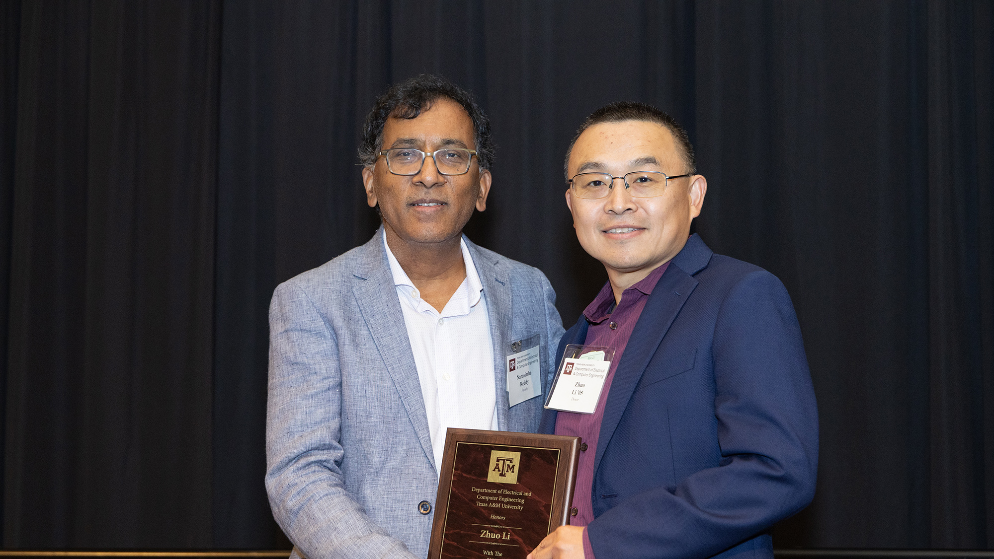 Two men hold an award, smiling and standing in front of black curtains.