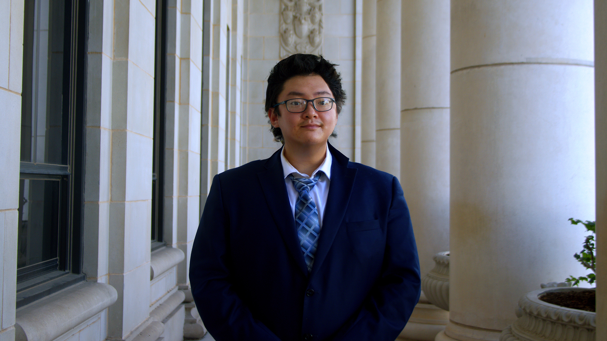 A man standing in a columned building entryway dressed in a blue suit.