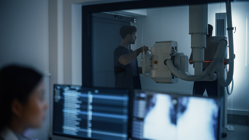 Male Nurse Assisting a Patient in Positioning Himself Before an Advanced X-ray Machine, Ensuring a Precise Procedure. Behind the Window, Female Radiologist Analyzing Body Scans on a Computer Monitor