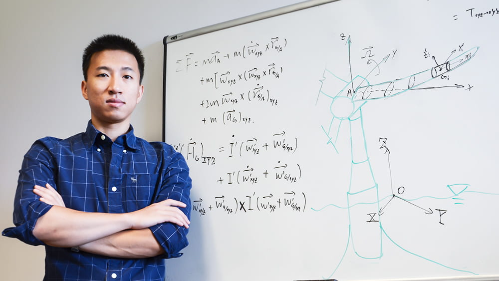 A graduate student stands in front of a dry-erase board of equations and drawings representing his research with wind turbines in the ocean.