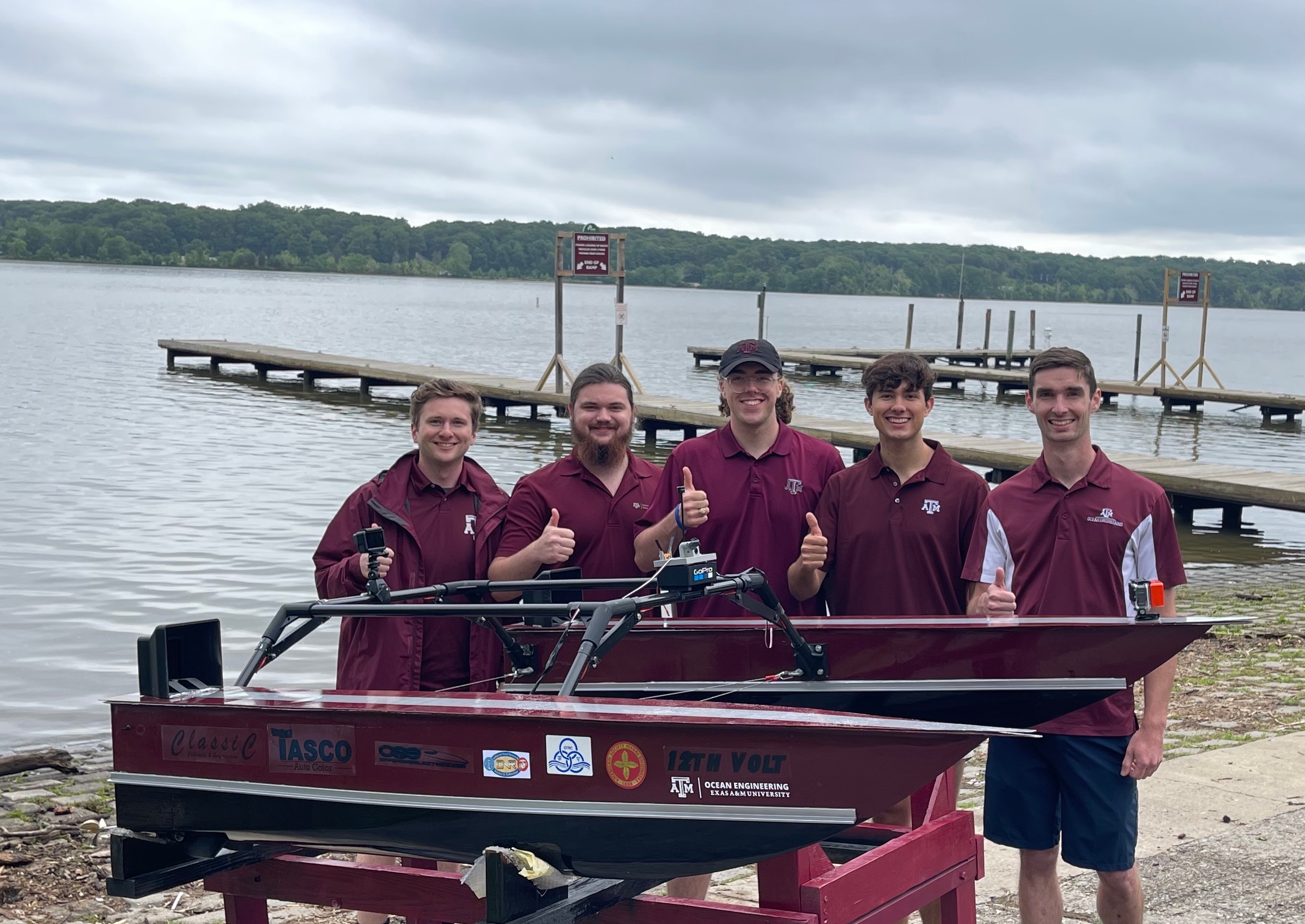 A group of students in A&amp;M polo shirts pose with thumbs up by their electric boat on the lakeshore. 