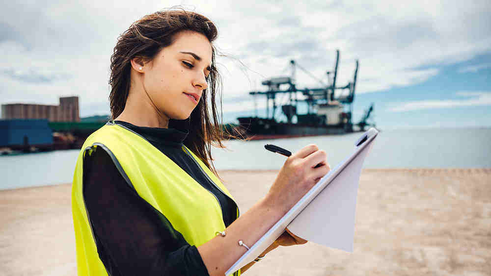 Female engineer makes notes on blueprints while standing near shore with an oil platform in the background.
