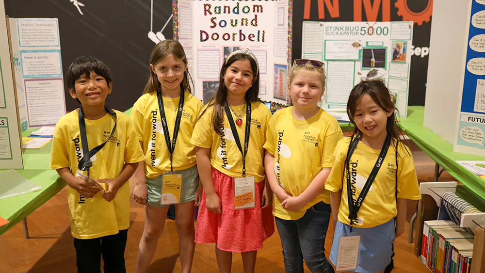 Five children wearing yellow shirts stand in front of a project board smiling at the camera. 