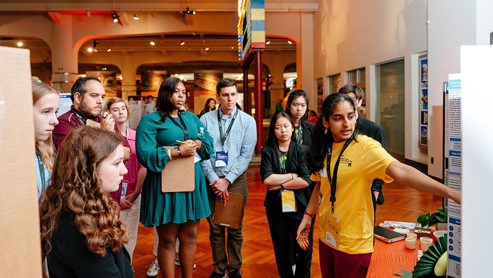 A female student wearing a yellow t-shirt presents her project to a group of adults and middle school students standing in a semi-circle in front of her. 