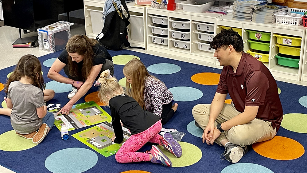 Two Spark instructors sit on classroom floor with three elementary students working on programming small robots to move along map path. 