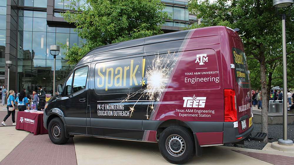 Maroon and black van with word Spark in Yellow sits in the courtyard in front of a glass building. 