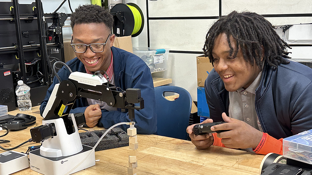 Two male high school students smile while working with a robotic arm to pick up and stack cubes. 