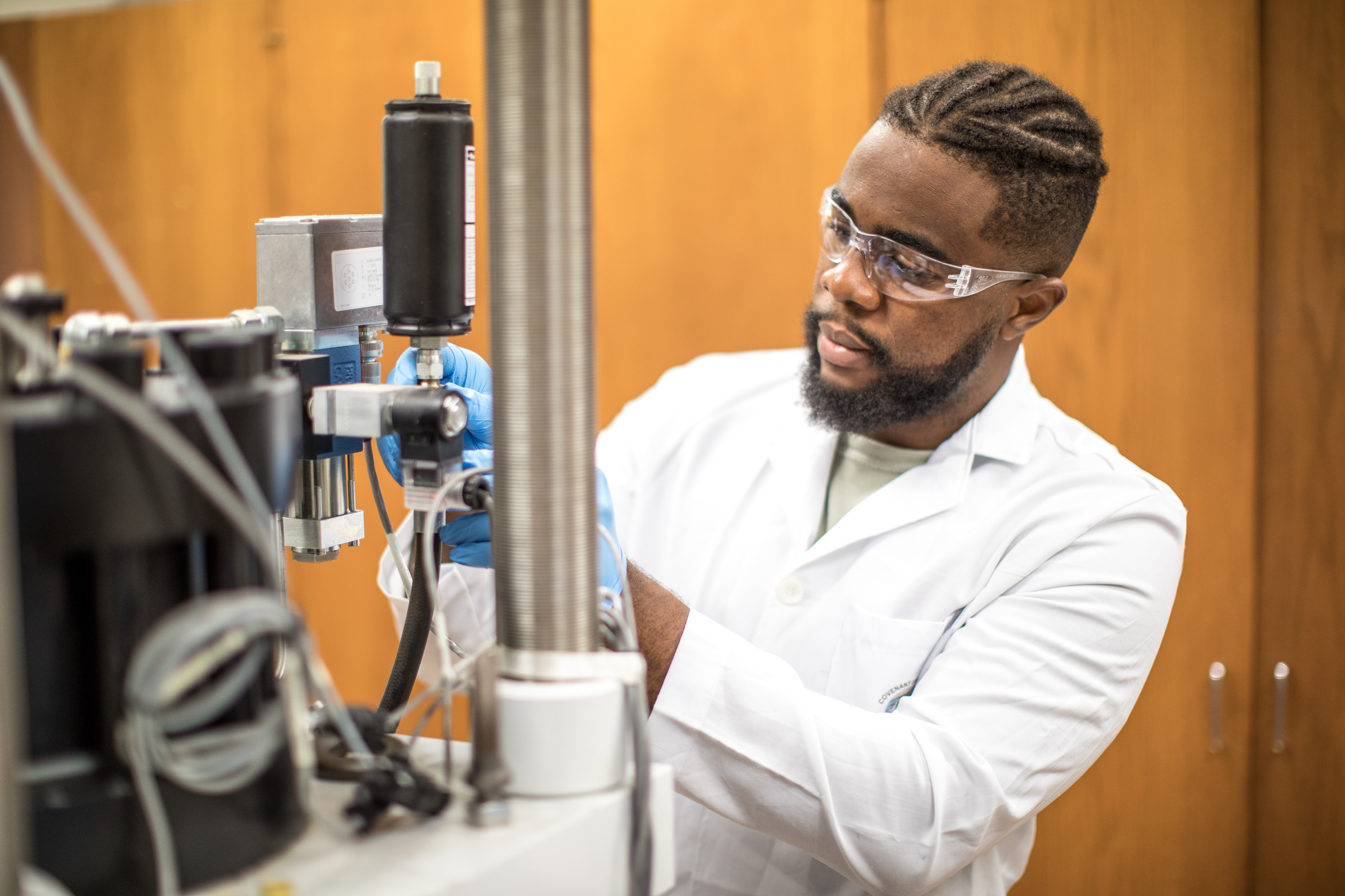 A man stands in a lab in a white lab coat. He has on PPE and is working with the lab equipment. 