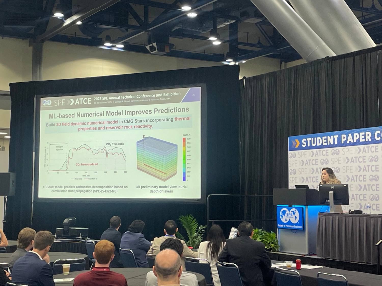 A woman stands on a stage with a large powerpoint on the screen behind her that she is presenting from. 