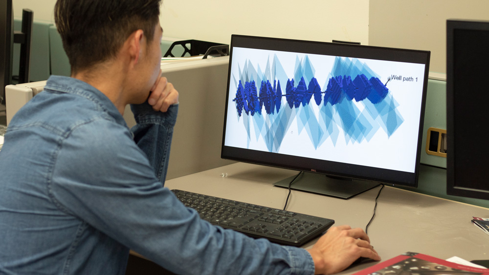 seated male petroleum engineering student at Texas A&amp;M University looking at a computer monitor showing a figure with a long black line, representing a well path, intersecting small and thick or long and thin angled planes representing two different types of areas within a reservoir