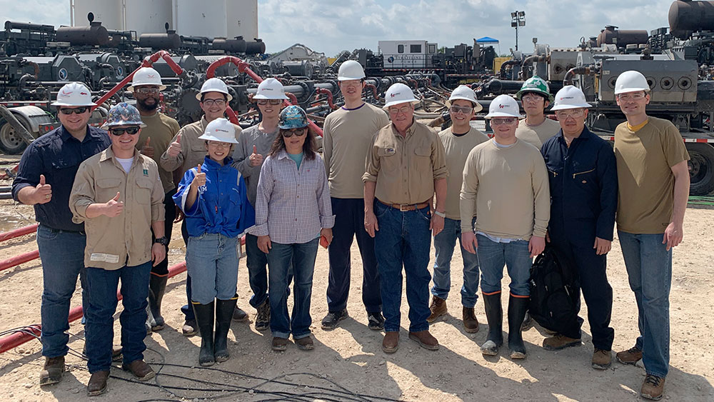 A dozen graduate students and two professors from Texas A&amp;M University standing outdoors on the graveled area of a well site surrounded by fracturing equipment, hoses and tanks