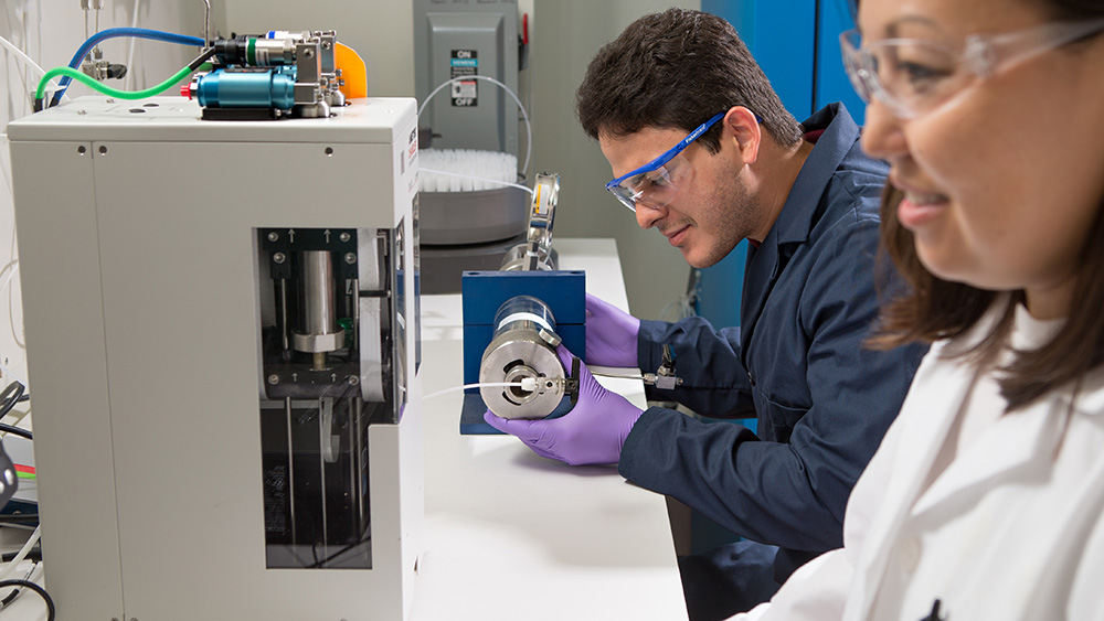 male petroleum engineering student researcher in protective attire inspects a piece of equipment while seated at a lab table as a female colleague stands nearby