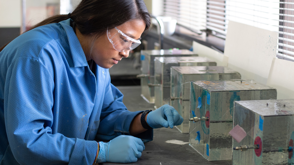 female petroleum engineering student at Texas A&amp;M University checking a clear polymer thread coming from a large cube of clear acrylic-type material approximately 12 inches long on each side with a small metal pipe running through it horizontally