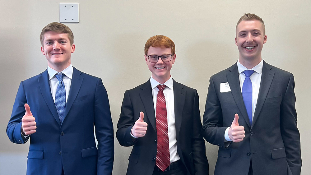 Three junior-level college students in business attire standing in a row and smiling.