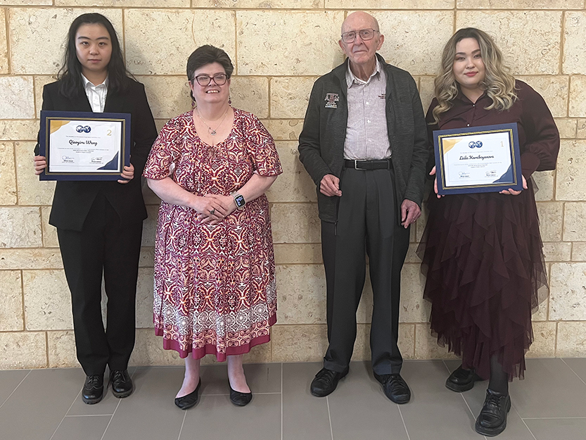 Two nicely-dressed doctoral students holding certificates and two faculty members from the Harold Vance Department of Petroleum Engineering at Texas A&M University stand in front of a wall.