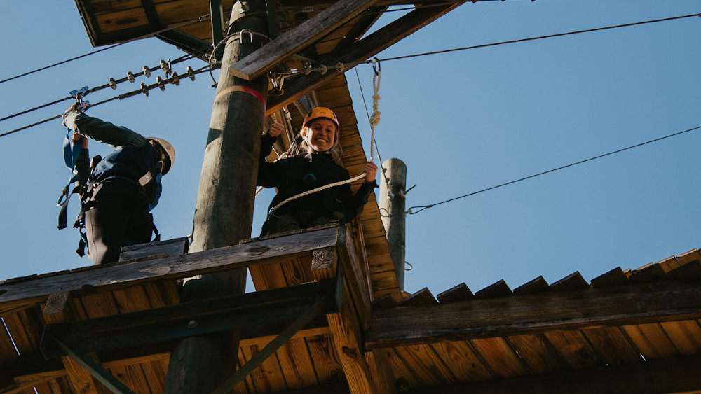 A young woman is at the top of tower, smiling at the camera before she zip lines.