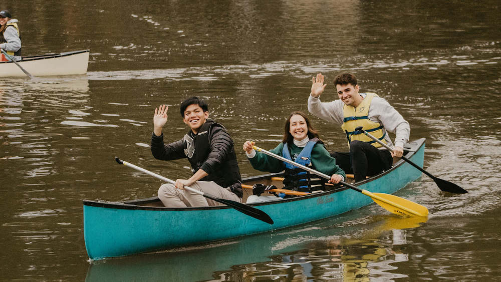 Three students are on a canoe on the lake waving to the camera.