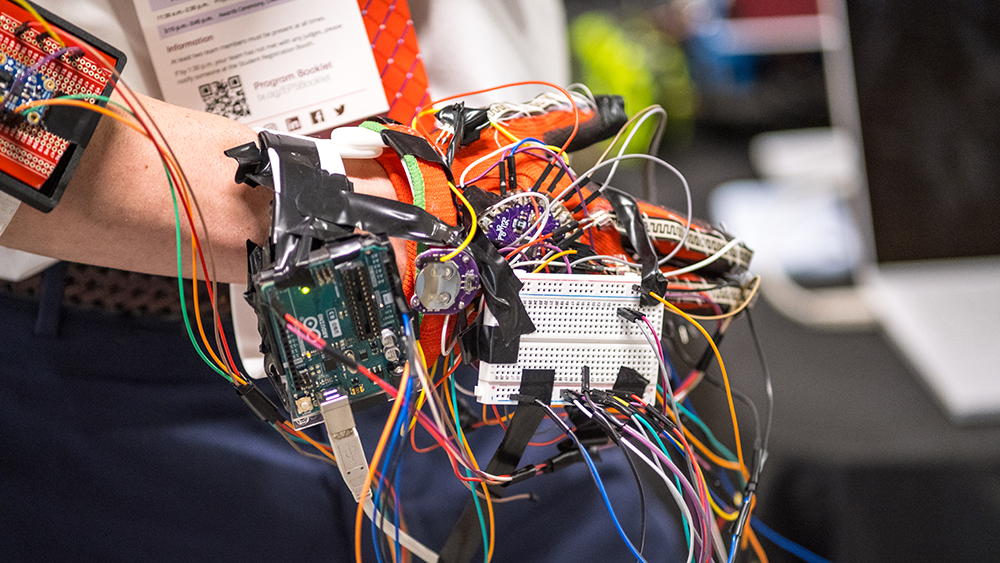 A close-up of someone wearing a red glove that has wires, microcontrollers, and a breadboard attached to it. It looks very complicated and DIY.